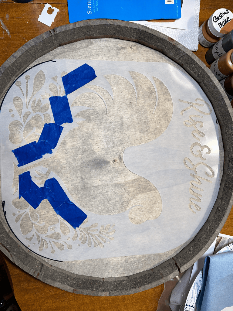 Reusable rooster stencil held in place with blue painter's tape on a wood round