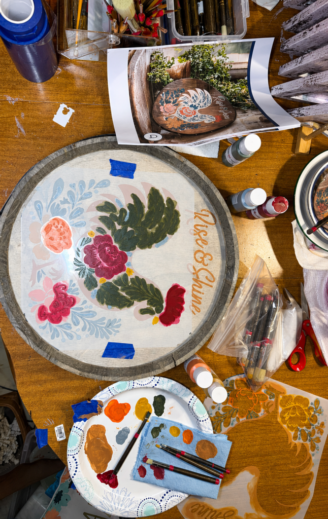 Hand-painting folk-art flowers around a chestnut rooster on a Rise and Shine sign