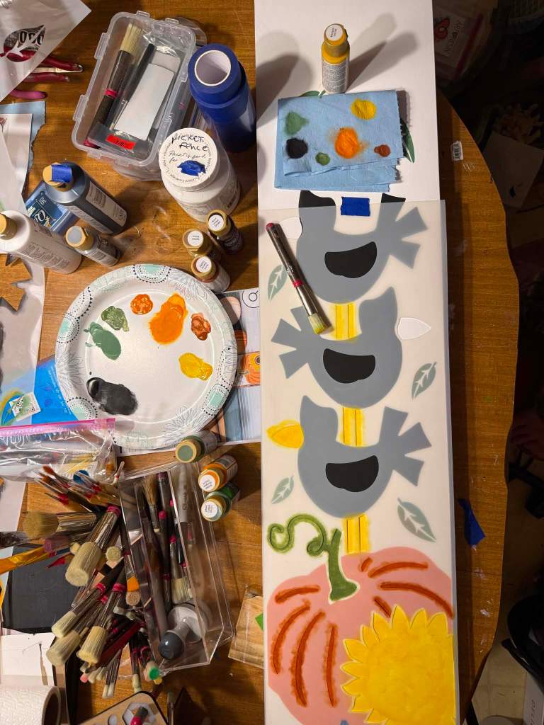 Overhead view of the crow stack porch sign in progress, with the pumpkin and sunflower basecoated and the crows painted in a gray underlayer