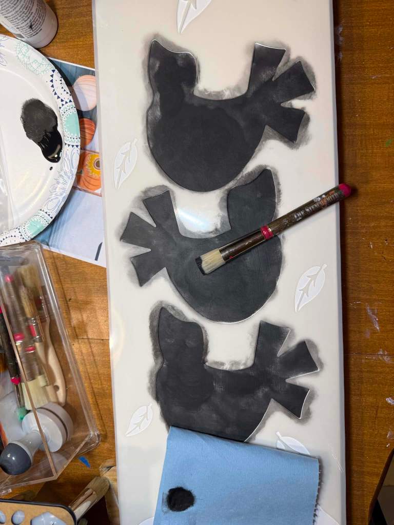 Painting the stacked crows black through the stencil using a nearly dry stencil brush, with paint offloaded onto a paper towel nearby