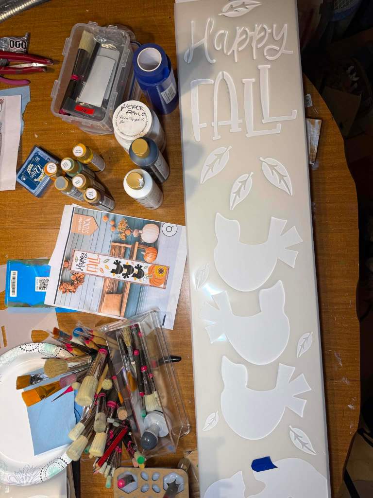 White mylar crow stack stencil taped in place on a primed porch board, with paint bottles, brushes, and a reference printout on the worktable .