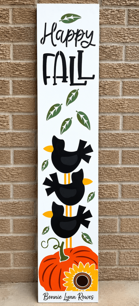 Hand-stenciled Happy Fall porch leaner sign with three stacked black crows, falling leaves, an orange pumpkin, and a sunflower, leaning against a brick wall 